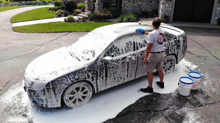 grey sedan covered with soapy water as a person holds a hose to rinse it